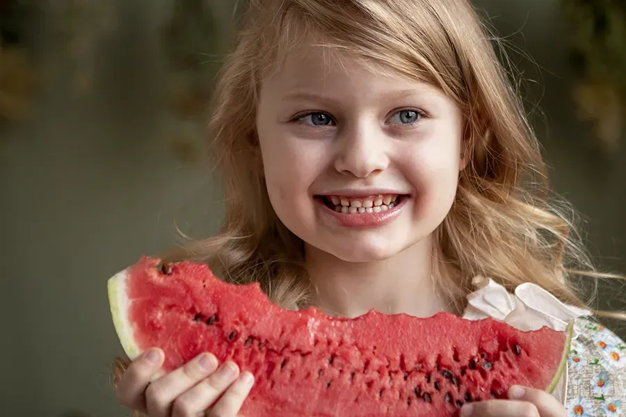 A little girl eating a slice of watermelon, illustrating the importance of healthy foods for children's dental health.